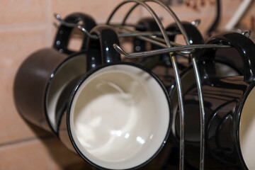 A set of stylish black mugs with white interiors is neatly arranged on a metal drying rack in a kitchen setting. The glossy ceramic reflection and functional dish storage create an atmosphere of home 