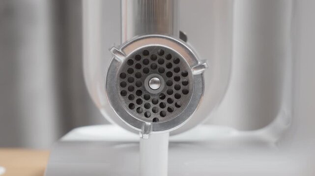 Electric meat grinder with loud motor sound. Metal mincer mechanism rotates rapidly behind perforated disc. Kitchen appliance working in empty state, steel blade spinning.