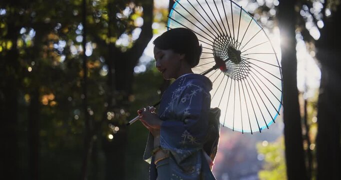 Locked off silhouette shot of Japanese woman in kimono standing in chilly autumn forest with umbrella