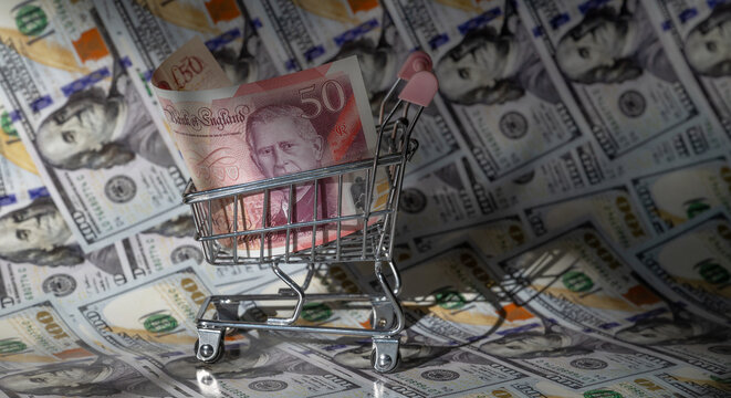 A 50 British pound note in a miniature shopping trolley with 100 US dollar notes in the background