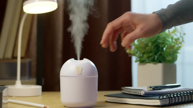 Human hand turns on white portable air humidifier on office desktop interior.