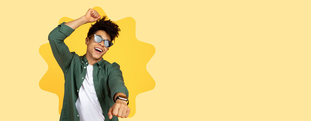 A young man is dancing joyfully in front of a bright yellow background. He is wearing sunglasses...