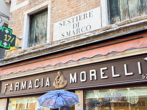 Venice, Italy - February 17, 2026: Farmacia Morelli storefront with a sign indicating Sestiere di S. Marco, featuring a decorative figure and an umbrella in front