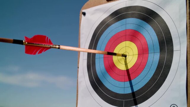 Arrow with red fletching hitting bullseye center of colorful archery target against blue sky, perfect accuracy and precision concept for goal achievement and success metaphor