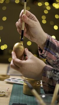 Female hands coloring eggs with golden paint on blurred garland lights