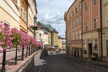 Fototapeta premium Vibrant cobblestone alley in Banska Stiavnica lined with pink flower baskets, pastel buildings, green roofed house, lamps and stone wall. Romantic old town vibe