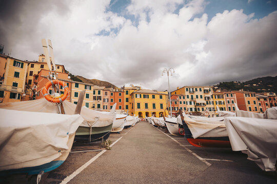 In a charming harbor, vibrant fishing boats rest under covers, surrounded by lively buildings. Dark clouds loom, hinting at an approaching storm, creating an exciting atmosphere.