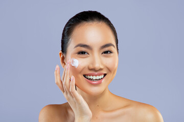 A woman smiles as she applies cream to her face in a plain indoor space during the daytime. She...