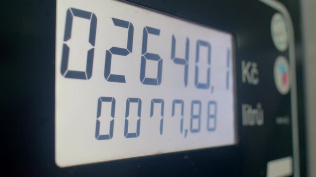 fuel pump display showing price totals, closeup lcd digits with koruna symbol, sales numbers rolling across screen, forecourt signage at edge, attendant absent, driver watches cost per liter, cashier