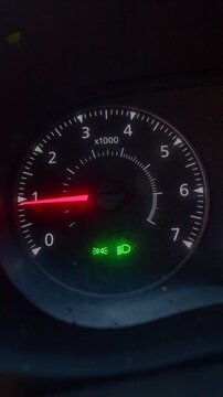 tachometer closeup at night showing glowing needle sweeping across dial near redline with green indicator illuminated, dark car interior, soft backlight and subtle grain, cinematic vertical framing,