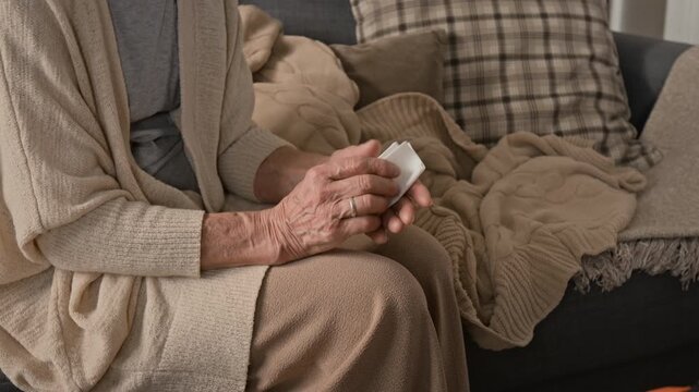 Midsection shot of anonymous elderly woman wearing beige cardigan fidgeting piece of tissue while sitting on couch at home