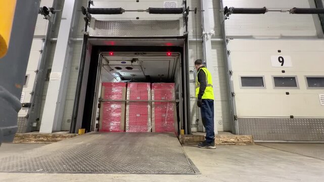 A warehouse worker in a high visibility vest stands next to an open loading bay door watching as pink shrink wrapped pallets are revealed inside a trailer