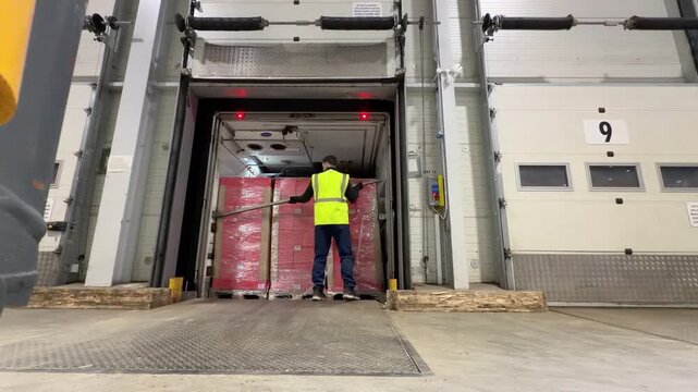 A warehouse worker in a high visibility vest enters the back of a semi trailer at loading dock number nine to remove a load bar before walking away