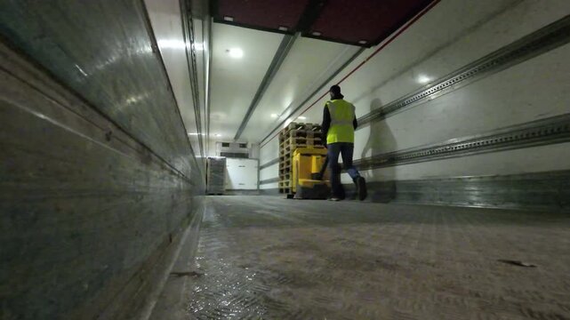 A low angle shot from deep inside a semi trailer as a warehouse worker in a high visibility vest pulls a yellow electric pallet jack loaded with empty wooden pallets toward the front of the cargo area