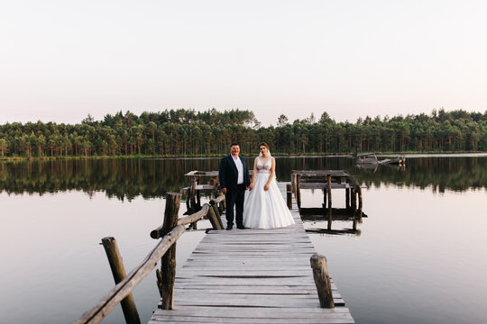 Wedding couple standing on rustic wooden pier over calm lake, surrounded by forest. Romantic landscape with reflections in still water, elegant bride and groom in evening light, serene outdoor ceremon