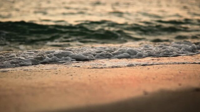 Soft sea waves with white foam washing over a sandy beach during golden sunset with beautiful orange reflections on the water surface in a peaceful tropical landscape with low angle view