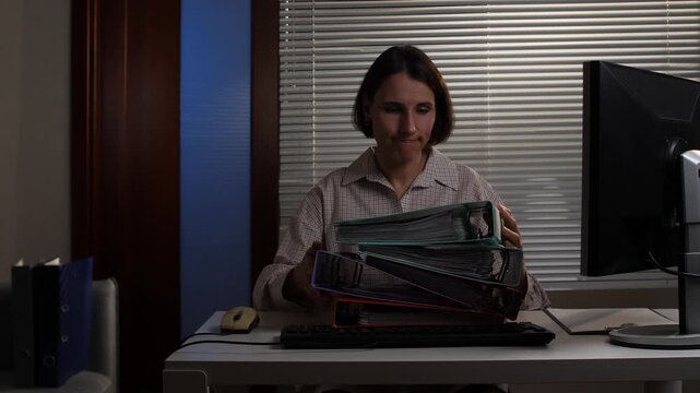 Focused female office worker sitting at her desk, carefully arranging a tall stack of files and binders, managing a heavy workload during an extended day