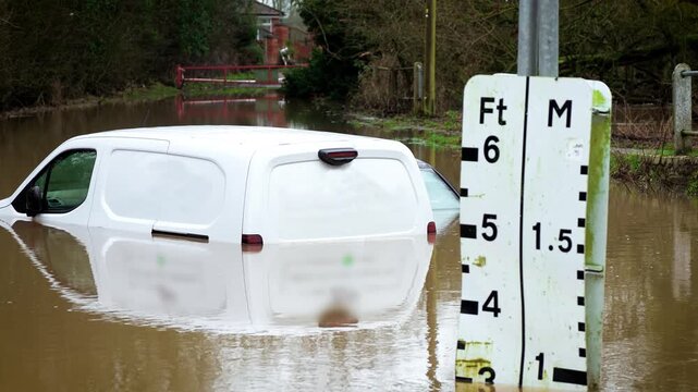Flood warning sign measuring water level while van remaining submerged on rural road. Brown river water rising and surpassing gauge markings during severe storm. Climate crisis worsening and causing