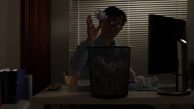 Overworked man sitting at his office desk, feeling stressed and frustrated with work while crumpling papers and throwing them into a full trash can in low light