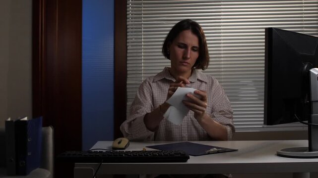 Woman sitting at a desk in an office late at night, tearing a document in half, expressing anger, frustration, and stress over work or a difficult personal situation