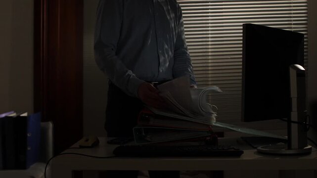Employee working late in a dimly lit office, standing at a desk with stacked binders and active computer monitor, secretly going through confidential files and paperwork
