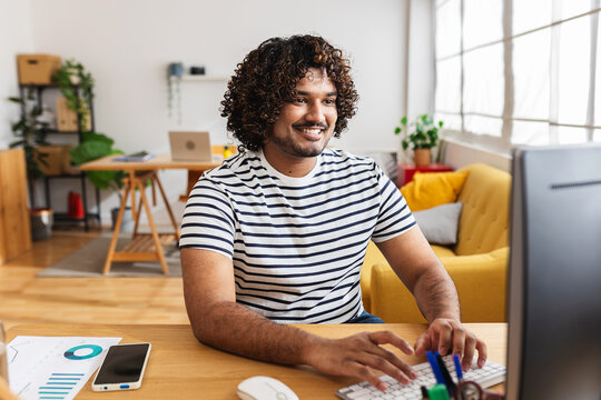 Young Indian man freelancer working on desktop computer at home office desk. Remote professional typing on keyboard and smiling while focusing on online project. Business and working at home concept.