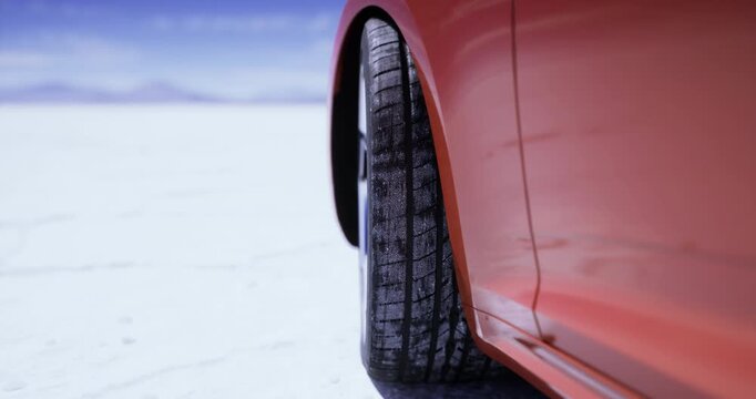 A bright orange car is positioned on a vast salt flat with a clear blue sky. The focus is on the tire, showcasing its tread pattern against the stark white landscape.