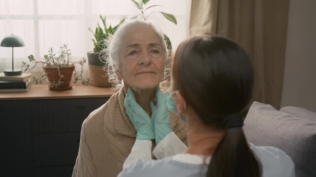 Female health worker using gentle circular motion to feel lymph nodes in elderly womans neck while carrying out home health check