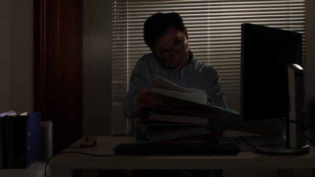 Overworked businessman managing multiple tasks, speaking on the phone while sorting through a large pile of documents at his desk in a dimly lit office late at night