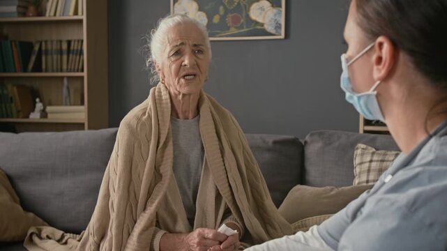 Tilt up rack focus view of young nurse wearing face mask unfastening orange bag and speaking with sick senior woman complaining of headache and coughing into piece of tissue