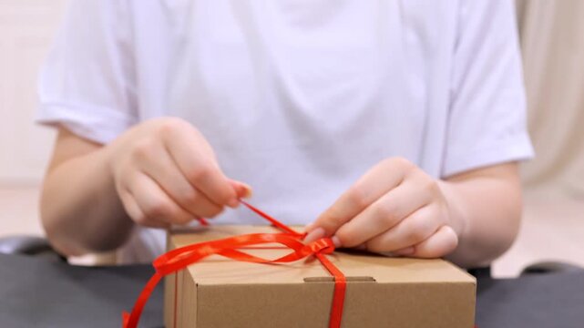 Person is tying a red ribbon around a brown box. The ribbon is tied in a bow