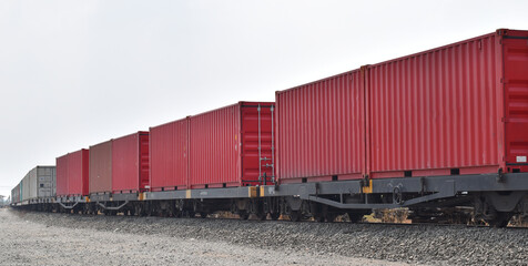Fototapeta premium Freight train transporting red shipping containers. Wide-angle shot of a long industrial cargo train on railway tracks under a clear sky, concept for global logistics, trade, and heavy transportation.