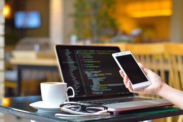 Full stack developer working in a cafe. Close-up of a programmer's hands holding a smartphone while coding on a laptop with a cup of coffee and glasses on the table, remote work and digital nomad. © Kosal