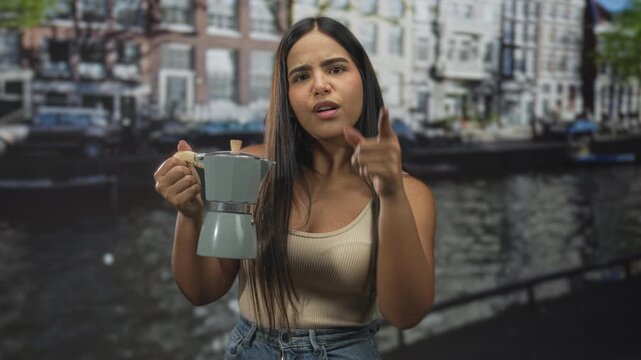 Young hispanic woman holds moka pot, points finger at camera with one hand and hand on hip on city street; morning ritual confidence.