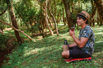 joven sonriente con sombrero sentado en cuclillas y bebiendo un mate en el parque. © Daniel