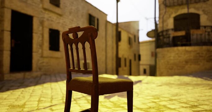 A wooden chair sits alone in a sunlit cobblestone street. Surrounding buildings feature historic stonework and inviting shadows, creating a tranquil atmosphere in a charming village.