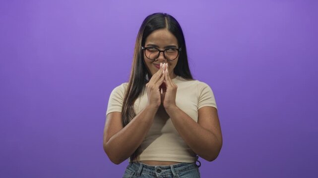 Young woman wearing glasses and a beige crop top clasping hands near mouth while smiling in a purple studio wall setting; playful mischief secret.