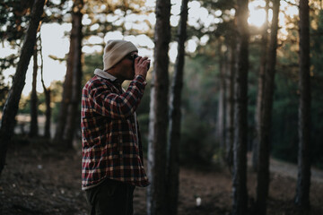 Fototapeta premium A man wearing a beanie and plaid flannel uses binoculars in a sunlit pine forest. He scans the wooded scene, capturing a calm outdoor moment.