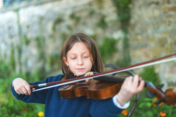 Young girl focused on playing the violin outdoors with a stone wall in the background