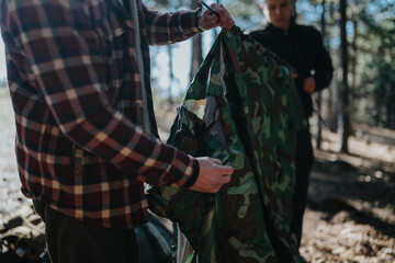 Fototapeta premium Two people in outdoor gear unfold and fold a camouflage tent in a wooded area. The scene captures a calm camping moment with natural light and trees in the background.