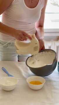 woman whipping cream with an electric mixer for meringue roll, berries and mint on table with baking ingredients. white tablecloth in soft natural sunlight, cozy home kitchen. vertical video, copy spa