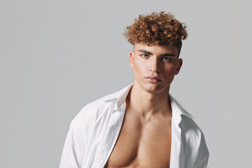 Obraz premium Young man with curly hair and open white shirt posing against gray background for fashion portrait and style concept with confident expression and casual look.