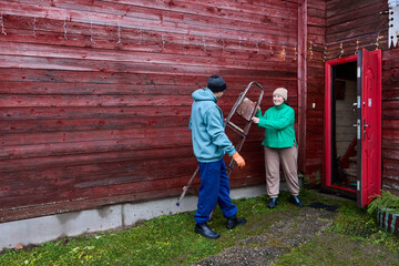 After hanging Christmas garland on house facade man carries ladder inside.