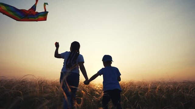 Two children holding hands in field. Children flying kite in field at sunset. A young girl and a boy are silhouetted outdoors for fun. A pair of children grasping each other's hands in a lifestyle.