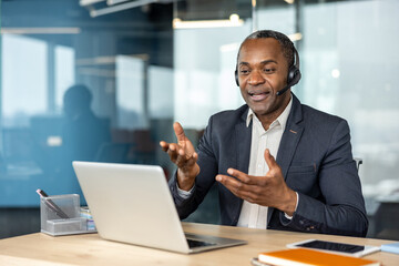 Fototapeta premium Mature businessman wearing a headset and suit jacket communicating on a laptop during an online video call from a modern office, actively discussing and gesturing