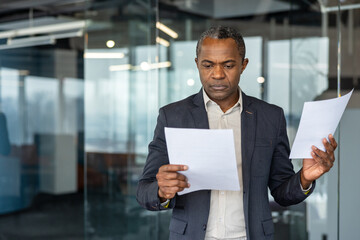 Serious african american businessman standing in a modern office, concentrating while reviewing...
