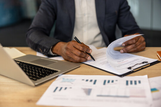 Person with dark skin tones signing a formal legal agreement on a desktop with a laptop and various white paper charts, symbolizing important corporate decisions and administrative tasks