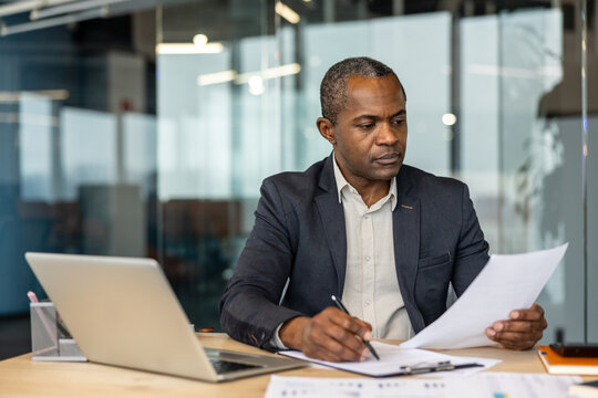 Mature afro executive concentrating on legal documents at his desk, reviewing contracts and making notes beside a laptop in a bright modern office, focused and professional