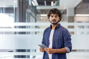 Naklejka premium Portrait of a young Indian man in a shirt standing in an office, holding a tablet and looking seriously at the camera