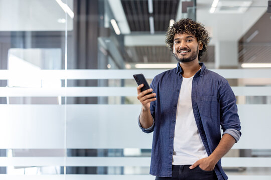Portrait of a young Indian smiling man standing indoors, holding a phone with his hand in his pocket and looking at the camera
